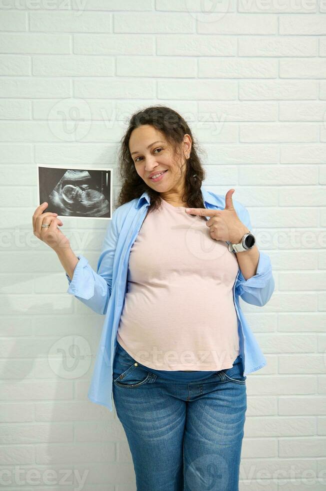 embarazada mujer sonrisas a el cámara y puntos hacia el ultrasonido a su bebé, aislado en contra un blanco ladrillo pared. foto