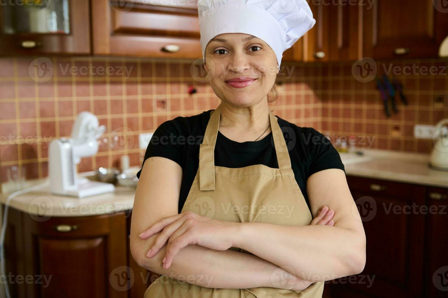 Pleasant housewife in chef's hat and apron, standing with arms crossed in the home kitchen, smiling, looking at camera photo