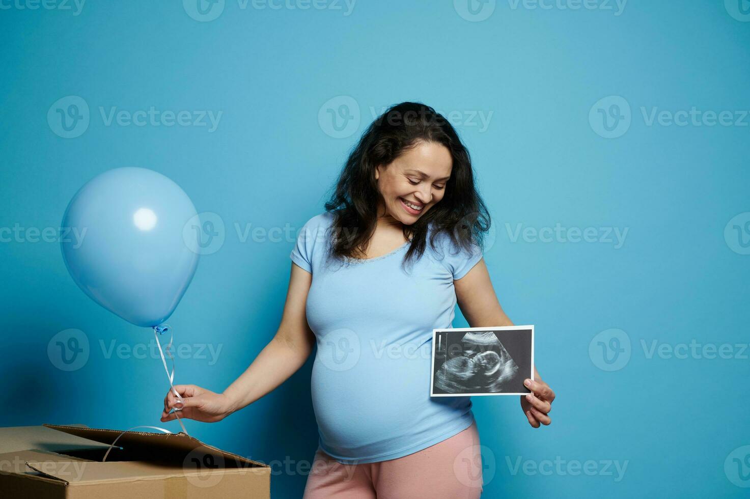 Beautiful expectant pregnant woman smiling, posing with blue balloon and baby ultrasound scan, isolated blue background photo