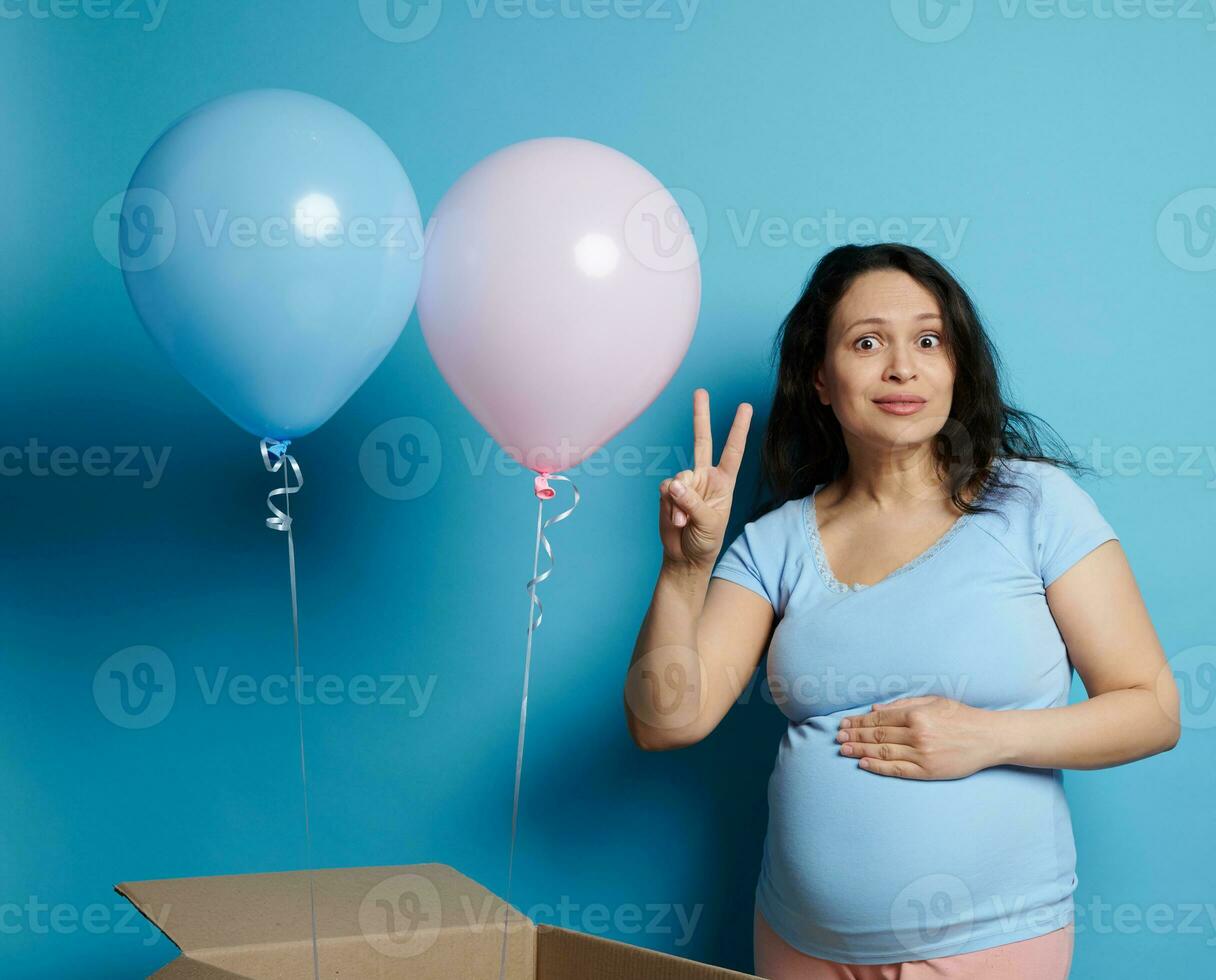 asombrado embarazada mujer esperando mellizos, posando con rosado y azul globos desde un caja durante bebé ducha o género fiesta foto