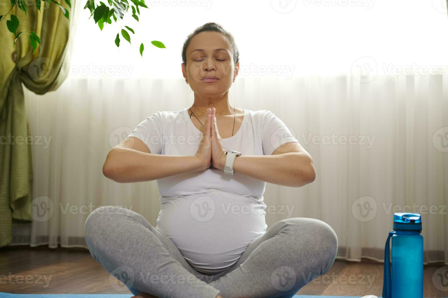 Pretty pregnant practicing some yoga at home, keeping hands palms together, closed eyes, meditating in lotus pose on mat photo