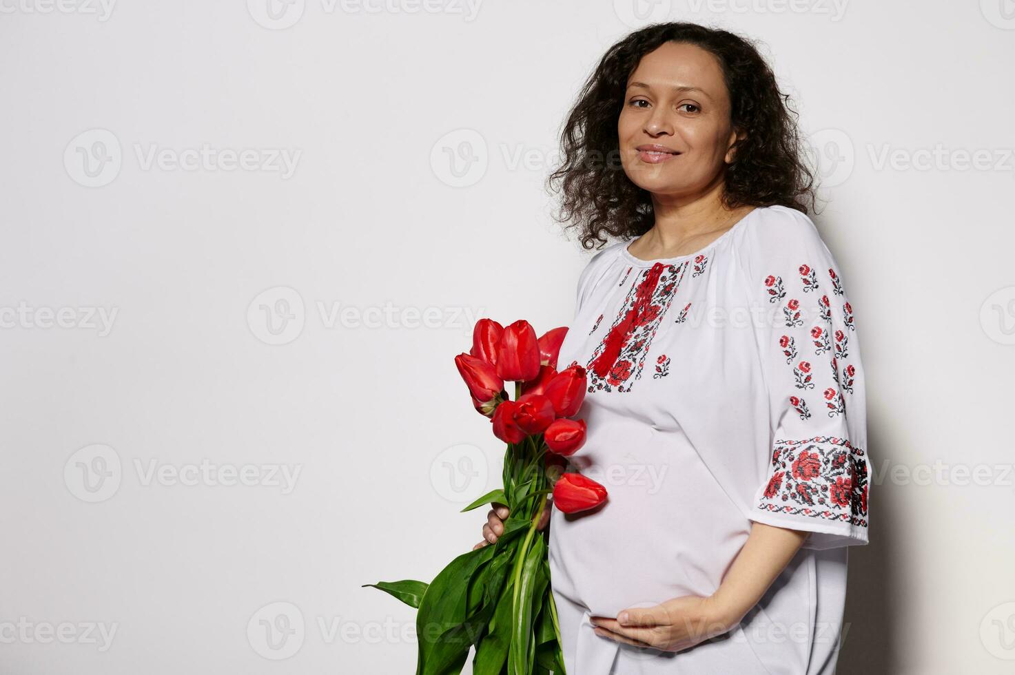 contento encantador embarazada mujer participación un ramo de flores de tulipanes, acariciando su barriga, sonriente a cámara en blanco antecedentes foto