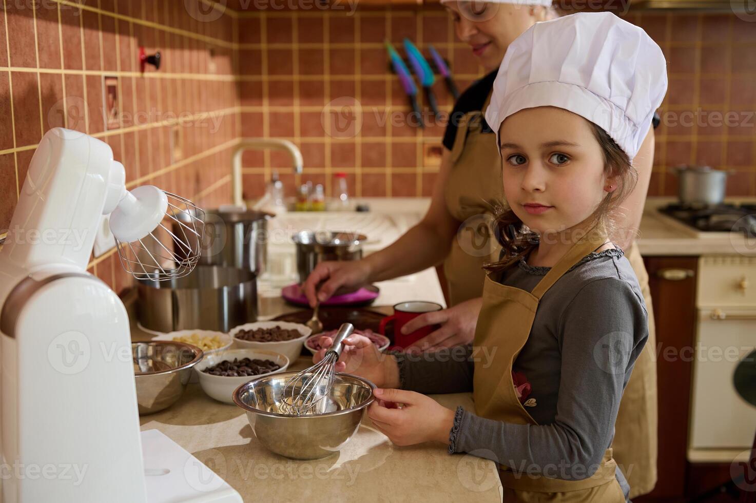 Lovely kid girl in chef's hat and apron, helps her mom to make a festive cake mixing whipped cream with melted chocolate photo