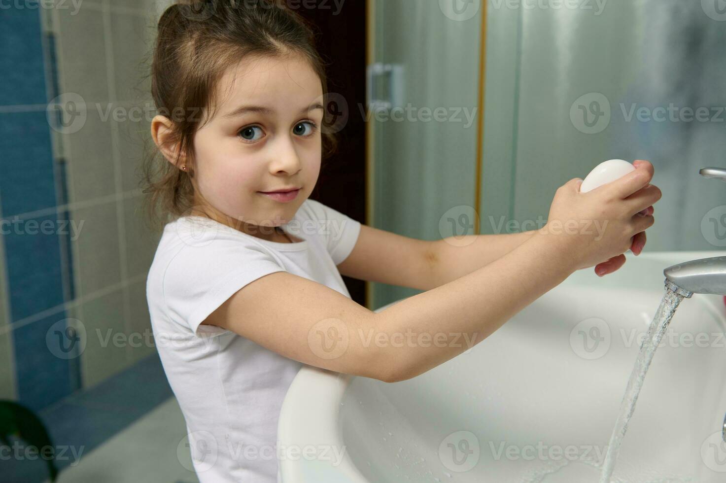 Beautiful little child girl smiling at camera, holding soap bar and washing her hands, standing by sink at home bathroom photo