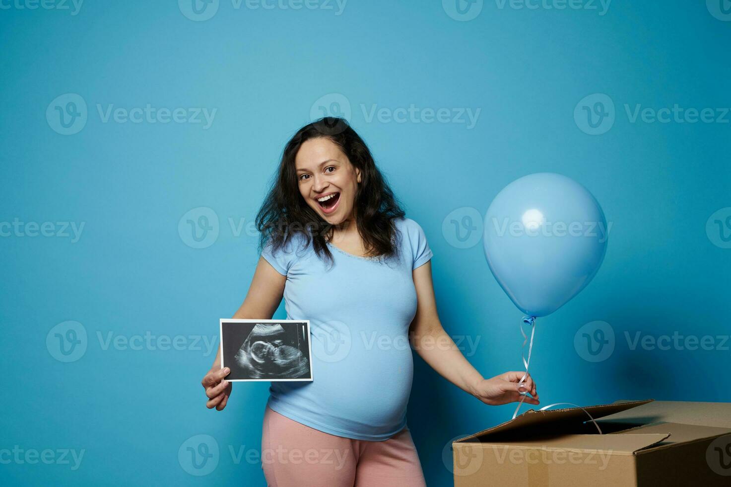 Happy cheerful pregnant woman smiling at camera, holding ultrasound scan image of her newborn baby boy. Gender party photo