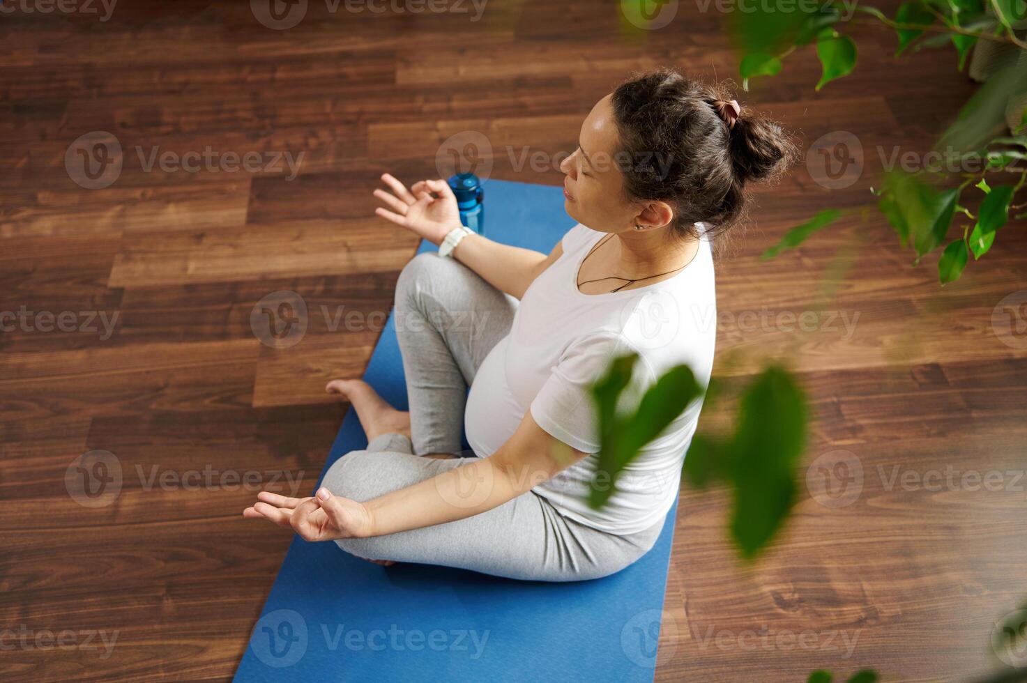 Latina pregnant woman sitting on mat in lotus posture, doing mudra sign with fingers, reaching zen, balance and harmony photo
