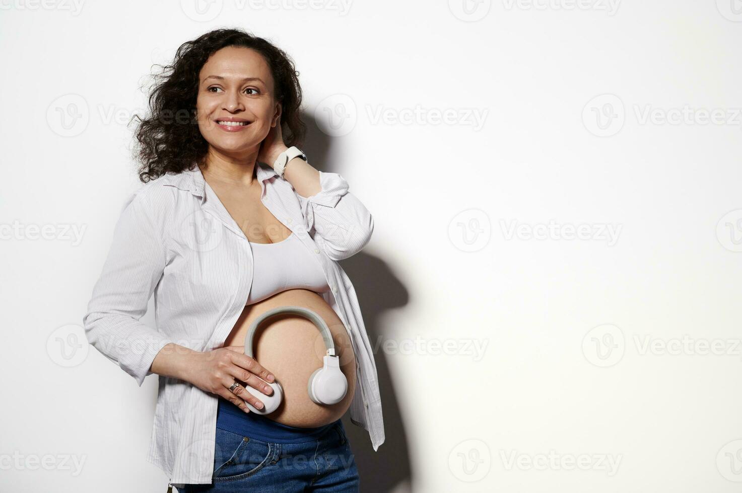 Stylish pregnant woman smiling looking away, putting headphones on her bare belly, isolated over white studio background photo