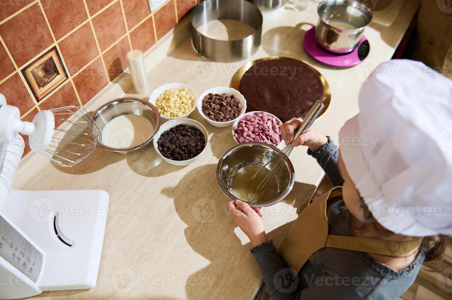 Overhead view of child girl confectioner using whisk, mixing ingredients in a metal bowl, standing by kitchen countertop photo