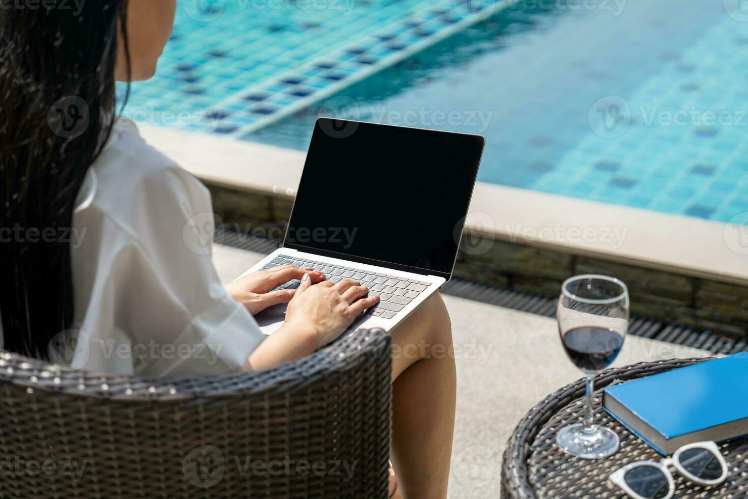 Young asian woman with long black hair is using laptop computer  with glass of red wine. She sitting at swimming pool photo