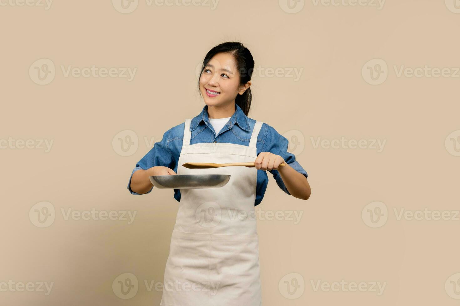 Portrait beautiful young asian woman wear apron with black pan and spatula on light brown isolated background photo