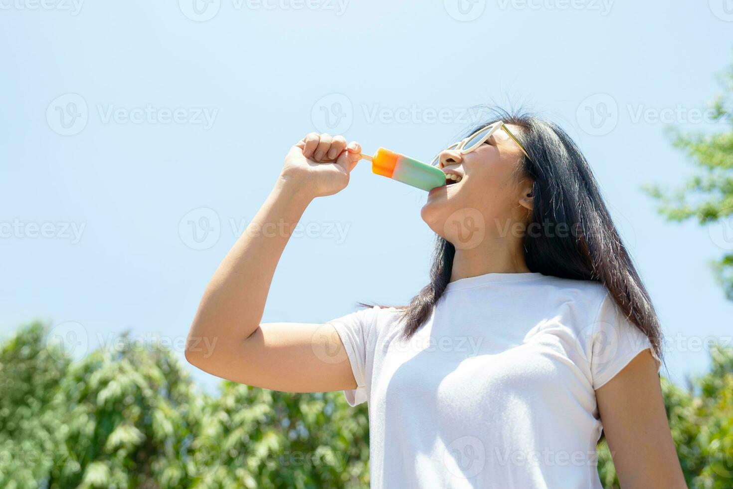 Smiling Asian woman wearing sunglasses holding green popsicle on blue sky background in summertime. Women eating popsicles, freshy summer season photo