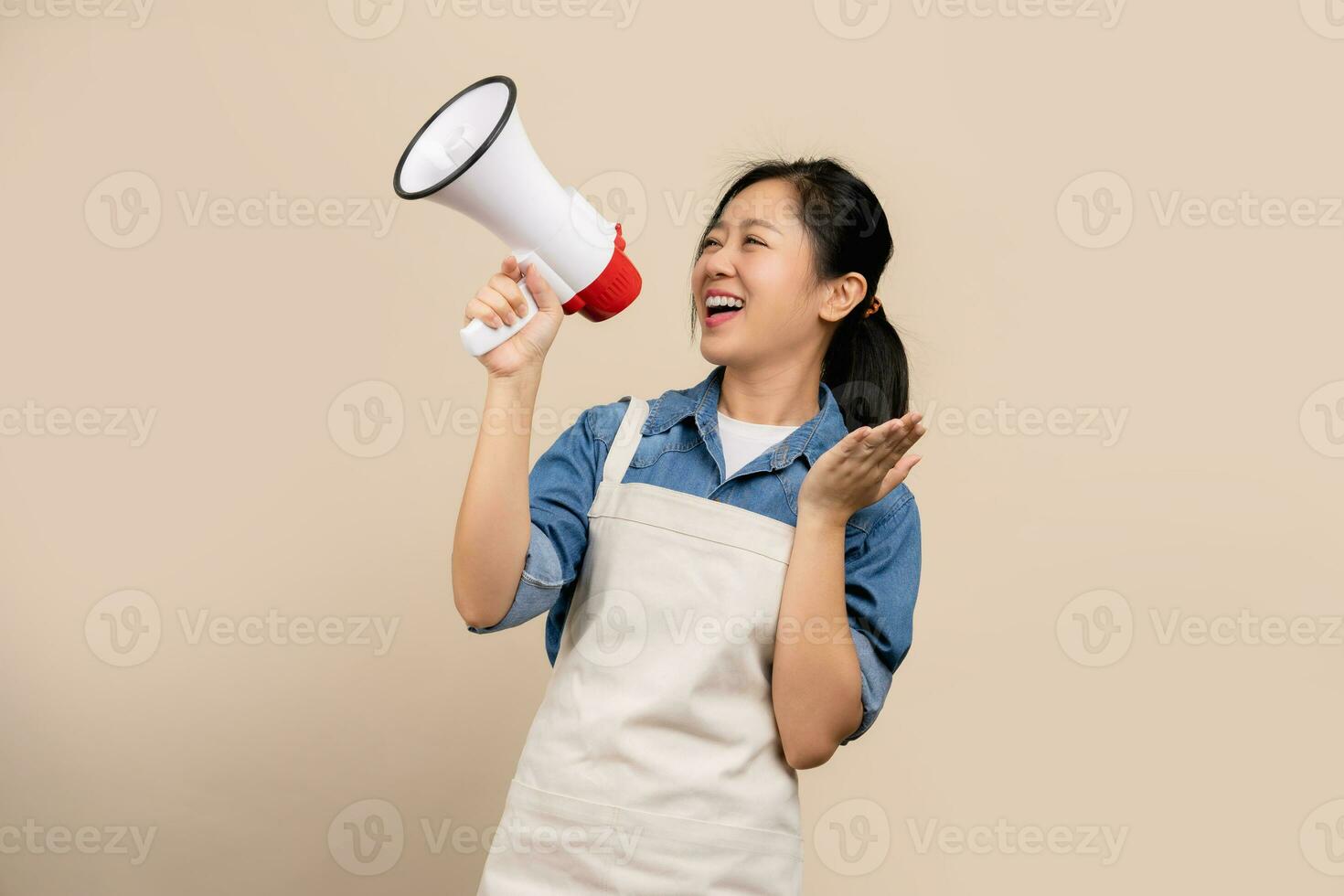 Beautiful Asian woman in apron with megaphone in hand. She is shouting excitedly standing isolated on a light brown background. photo