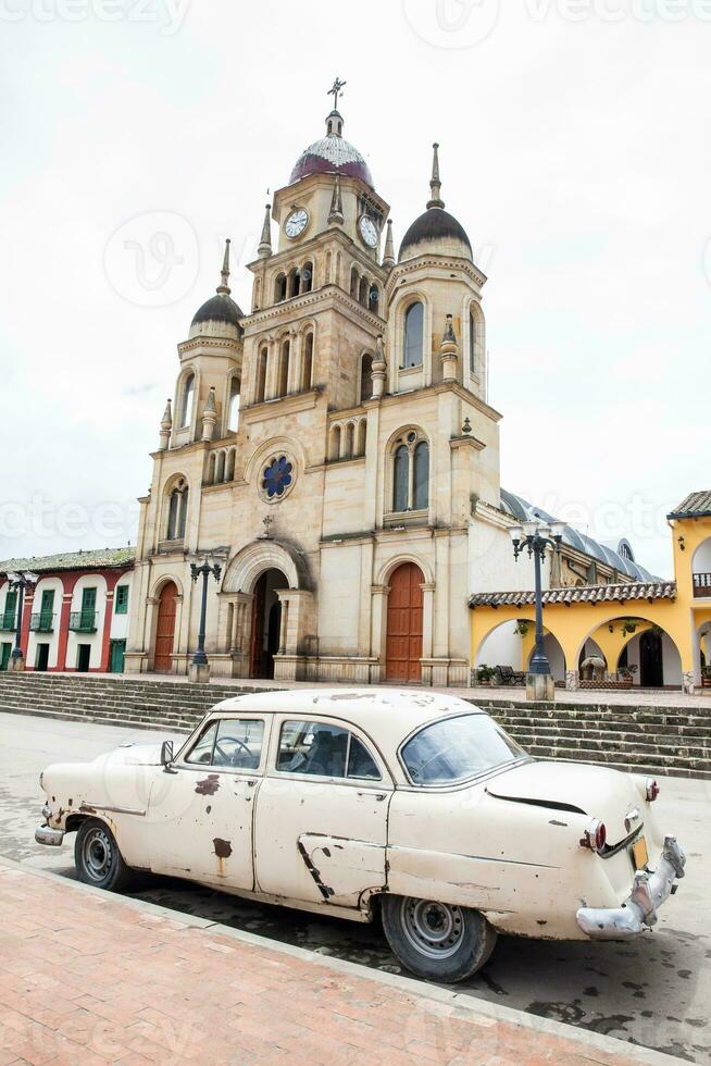 Antique rusty car parked next to the Parish Church of the small town of Ventaquemada in Colombia photo