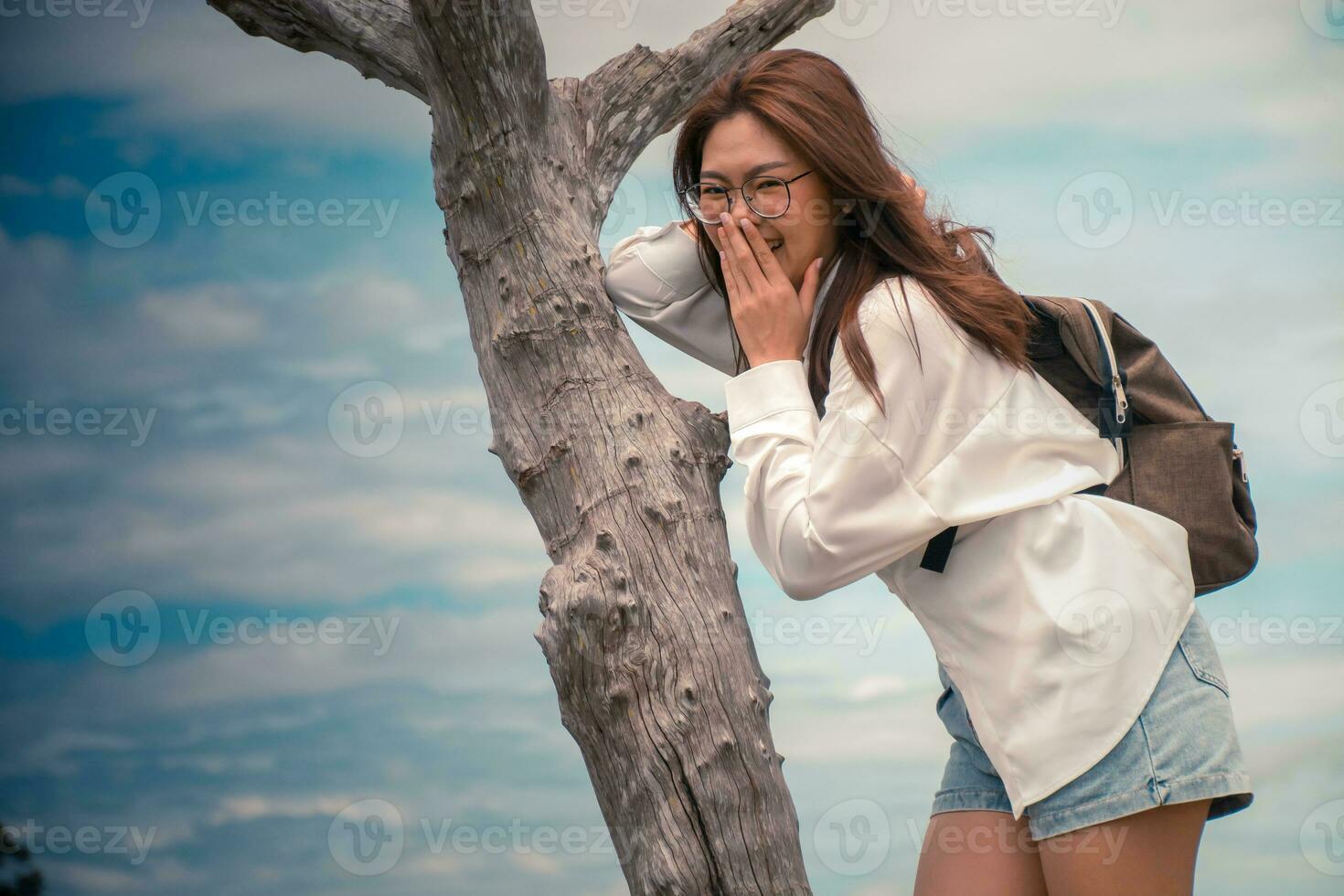 Beautiful asian woman wearing glasses and white shirt and jeans. standing with her right hand on a dead tree in background is clouds and sky. laughing, lifestyle, vacation life happiness concept. photo