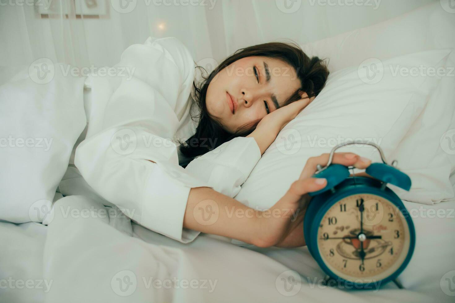 Asian beautiful woman in white pajamas turning off alarm clock while sleepy in bed at her bedroom of house or hotel on holidays. Girl is having trouble wake up late in morning. Unhealthy sleep. photo