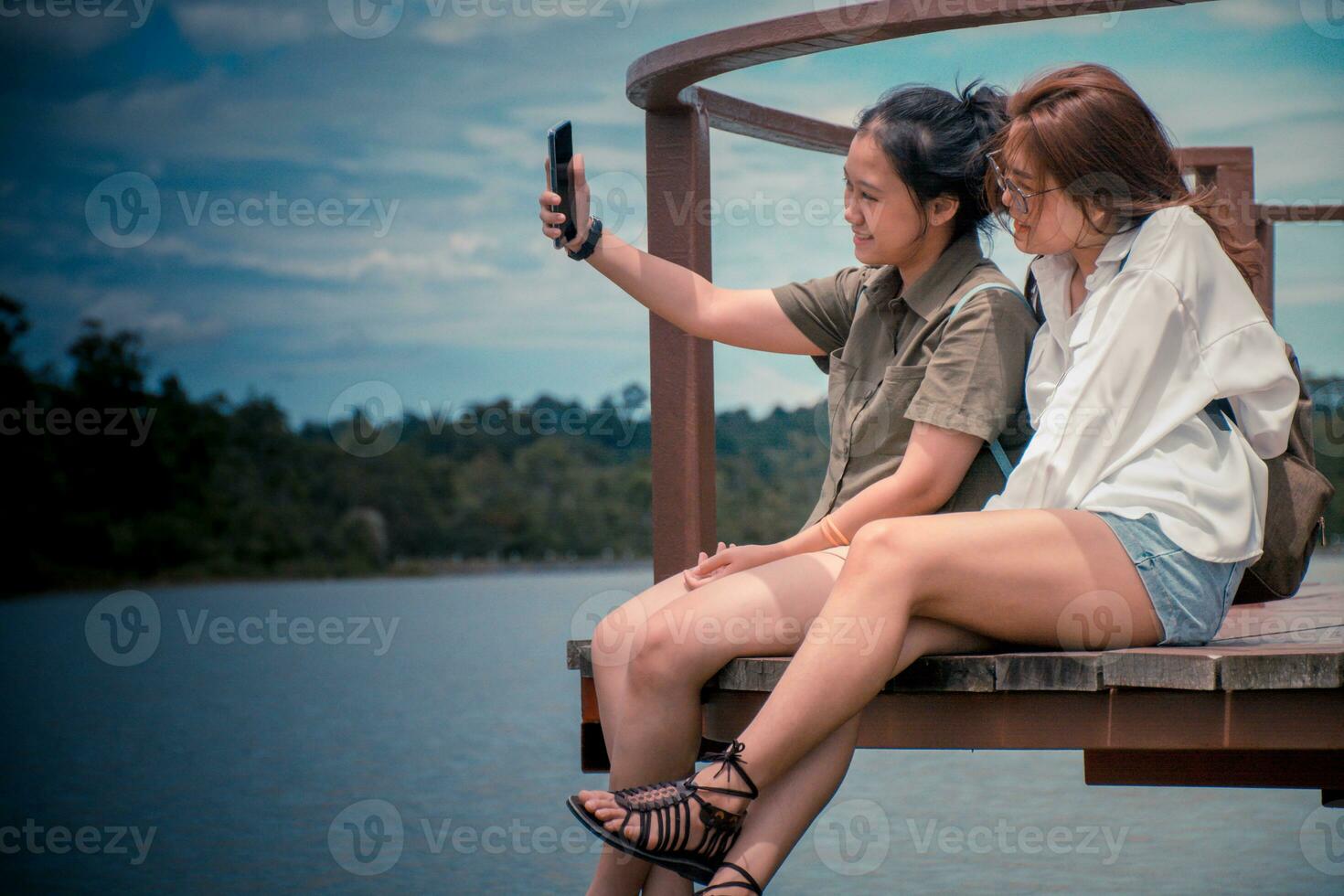 Two joyful cheerful girls taking a selfie while sitting together on the bridge and looking at the beautiful rivers and mountains. Two young asia womens backpack to travel the holidays. relaxed. photo