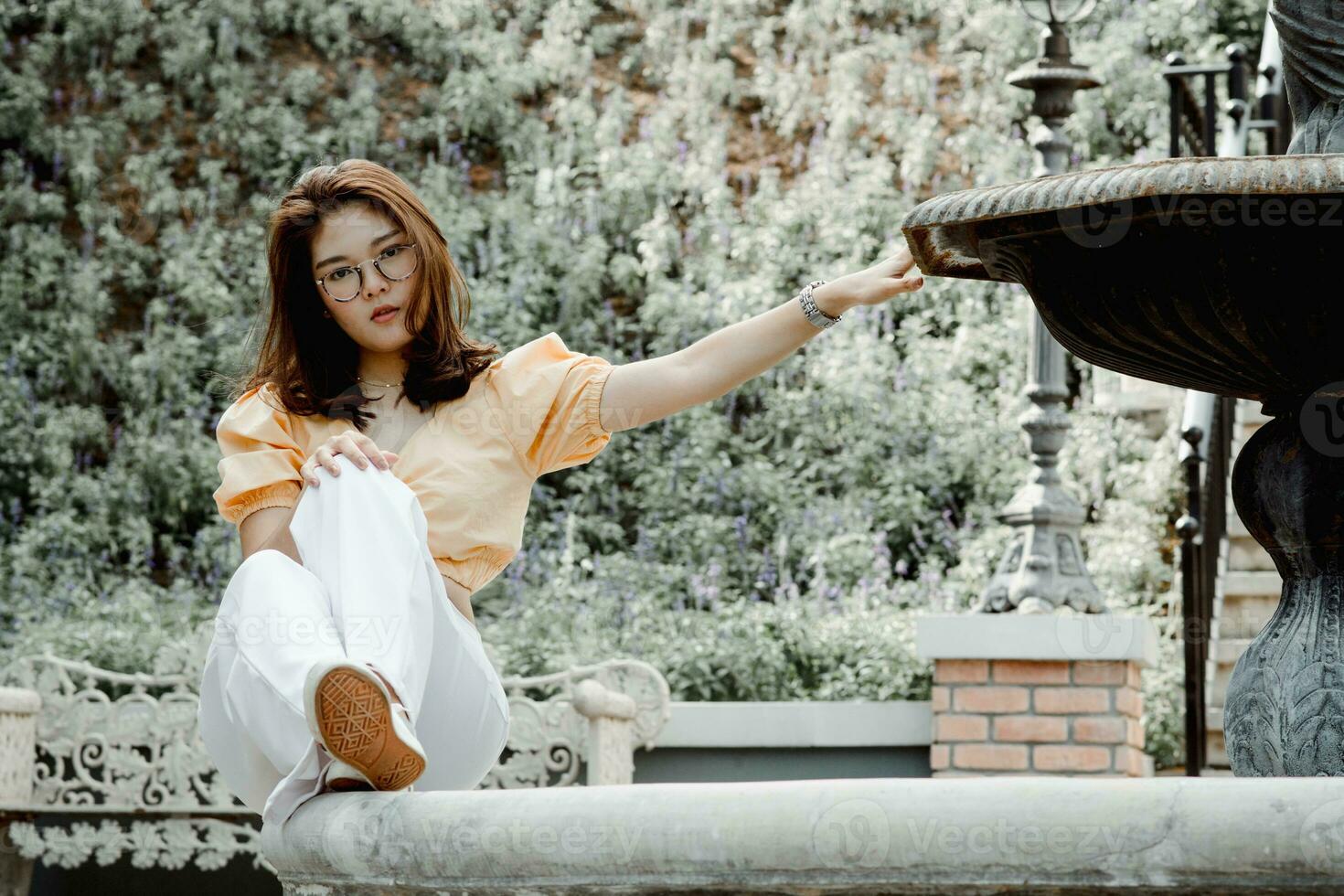 Beautiful young woman wearing glasses - yellow shirt and white pants. Travel on vacation. Posing for a photography, Sitting on the fountain, looking at the camera. The view behind is trees and nature. photo