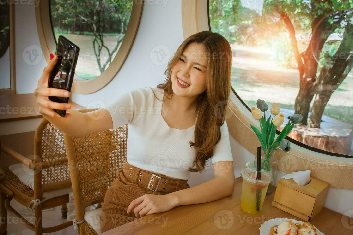 Portrait of cool cheerful girl having video call with lover holding smart phone in hand shooting selfie on front camera. Good mood lady with expansive smile enjoying started weekends in the cafe. photo