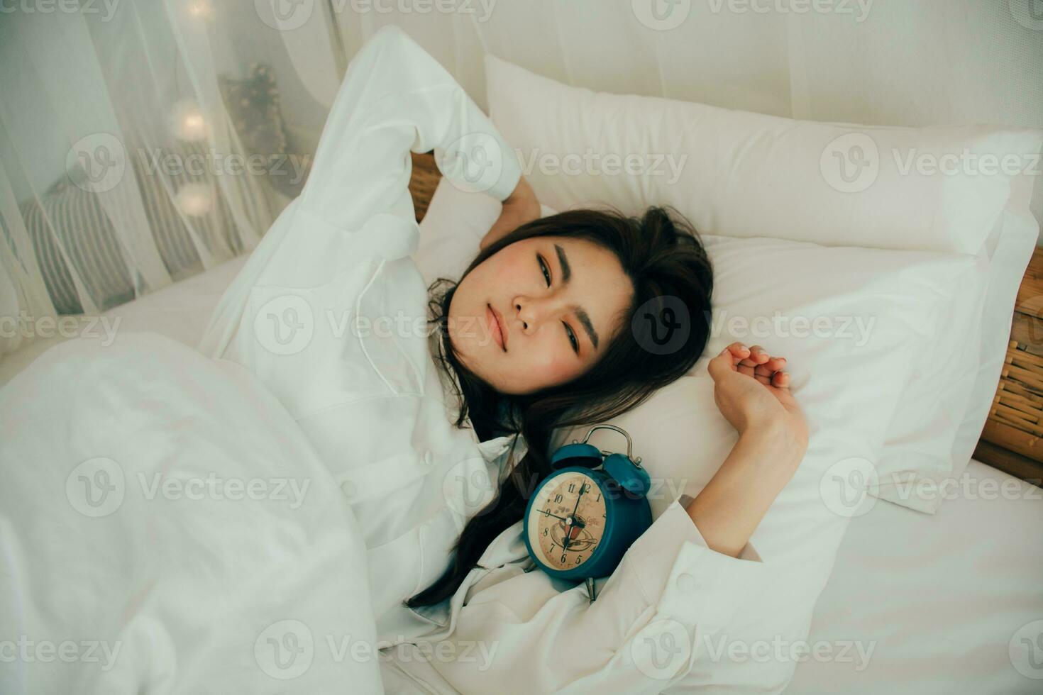 Portrait of attractive lovely girl enjoying time in bed after sleeping lying under blanket making stretching keeping eyes closed. Good day life health concept. Waking up in the morning. Green Clock. photo