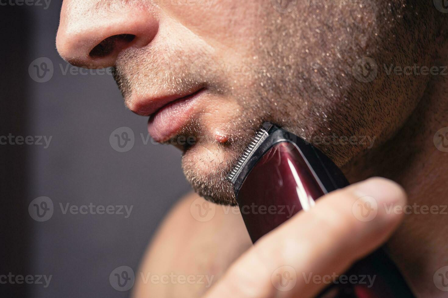 de cerca caucásico hombre afeitados su cara con un eléctrico recortadora cerca un Topo en su barbilla foto