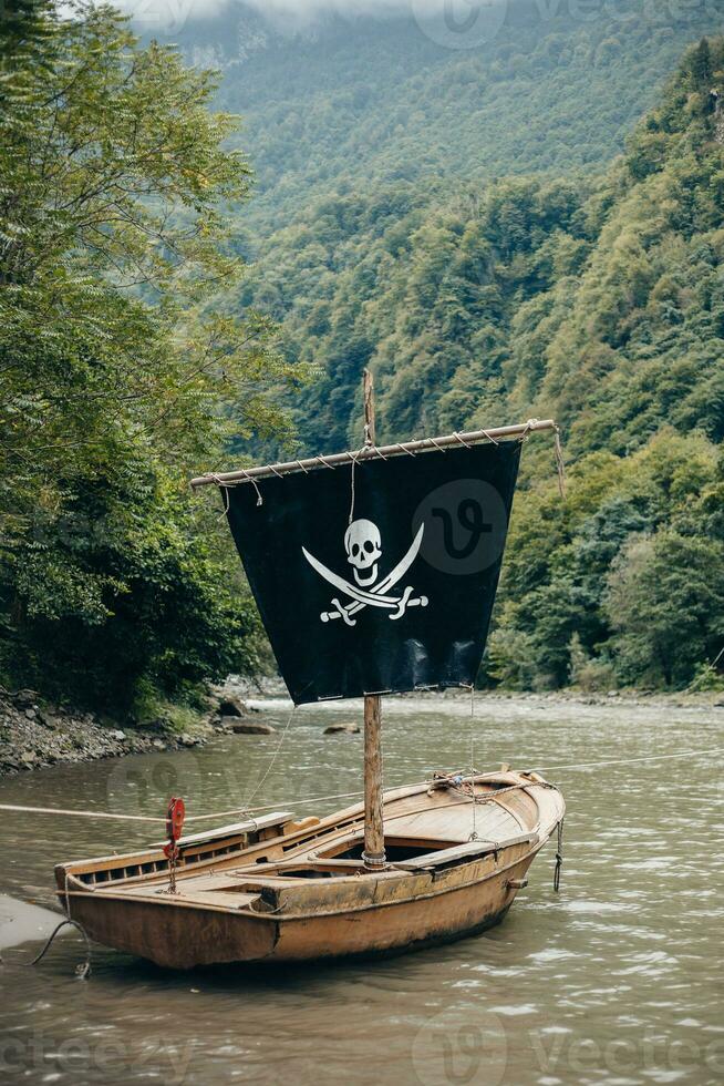 Pirate symbol on a black sail of a wooden boat near the river bank in the mountains - adventure in search of treasures - skull with bones jolly roger photo