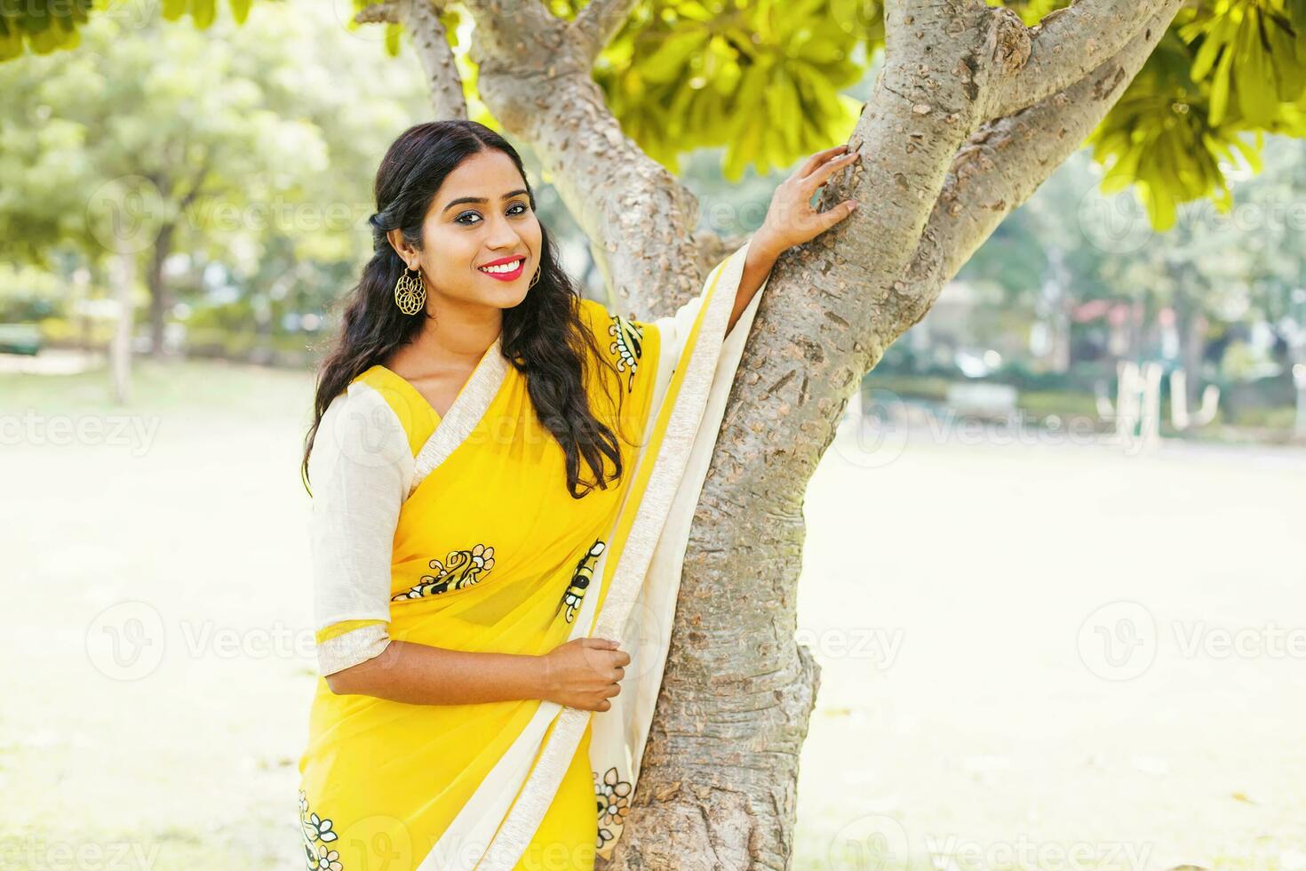 Young beautiful Indian woman in yellow chiffon saree posing near the tree in a park photo