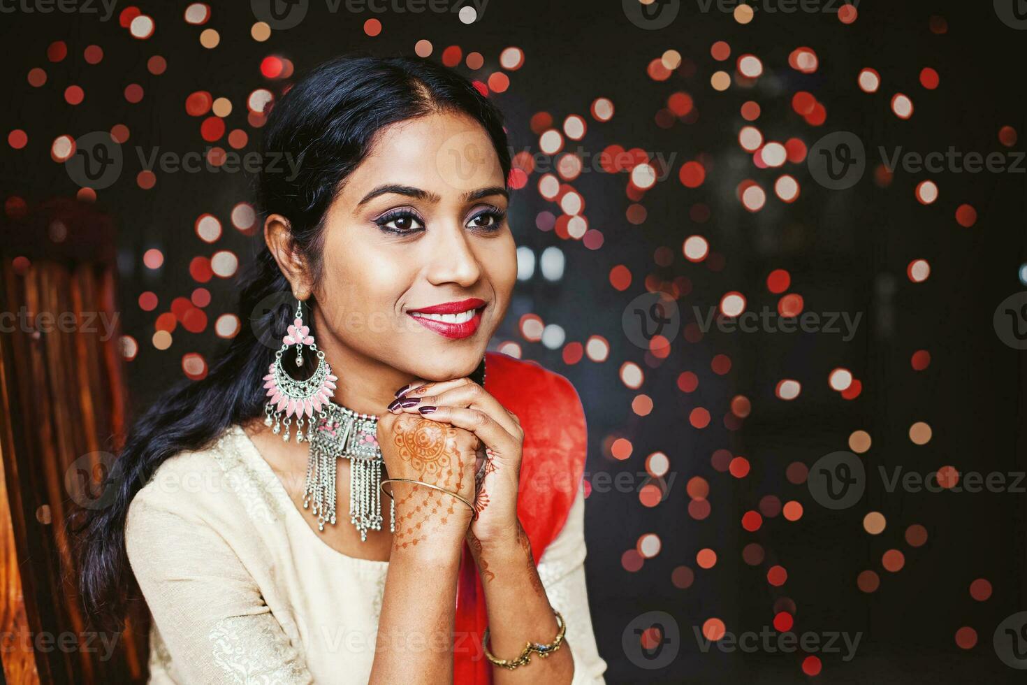 Young beautiful indian woman wearing jewelry and festive ethnic clothes sitting and waiting over bokeh background photo