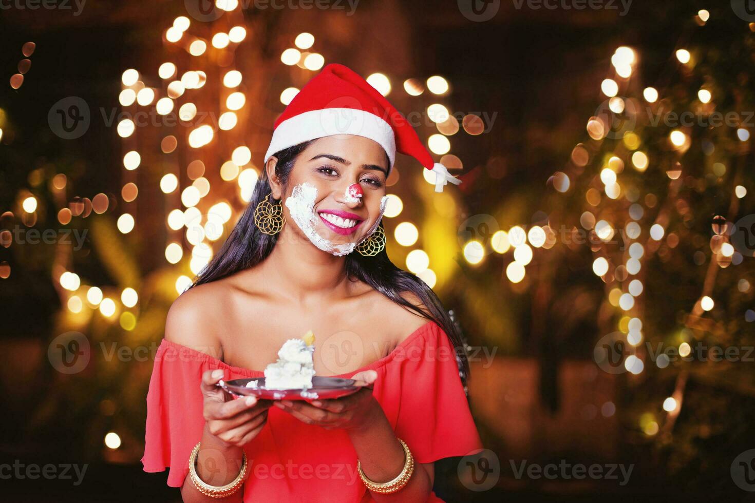 Pretty happy young Indian woman in New Year hat holding cake, cream smashed over her face photo