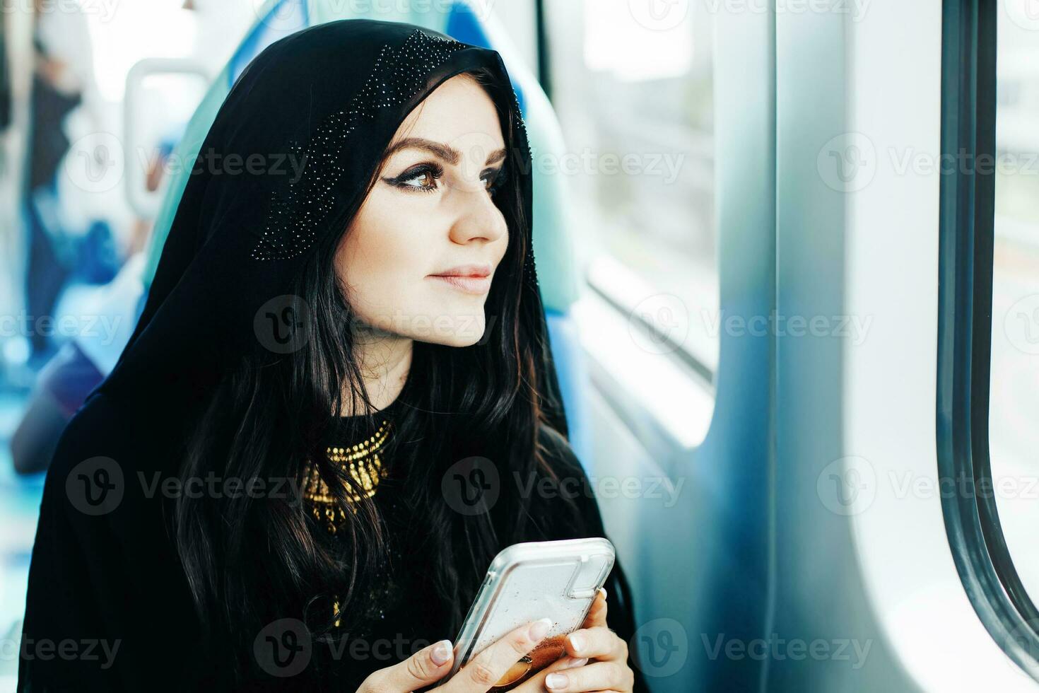 Beautiful Arab woman in hijab commuting in public transport, holding mobile phone, looking forward photo