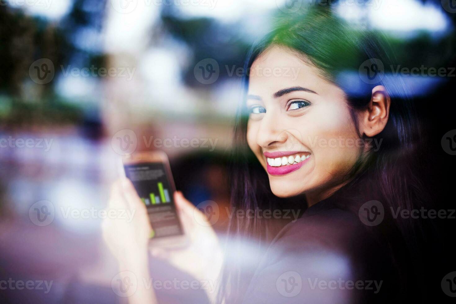 A beautiful young Indian woman smiling and looking through a glass window while holding a phone with graphs in her hands photo