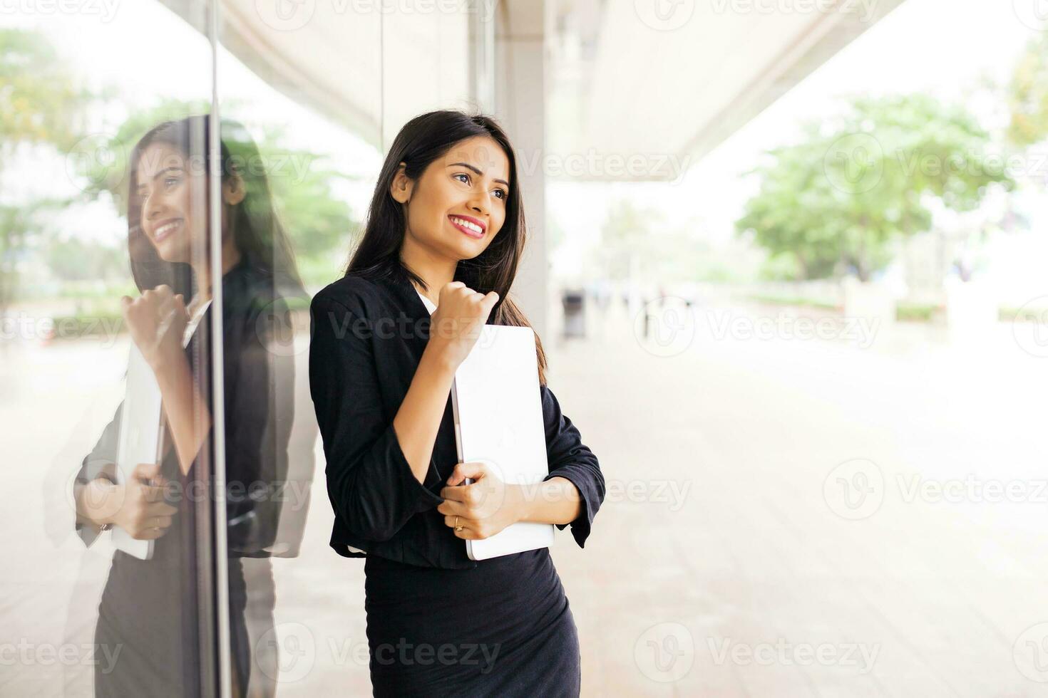 A beautiful professional Indian woman wearing a suit standing holding a laptop and smiling by a glass window photo