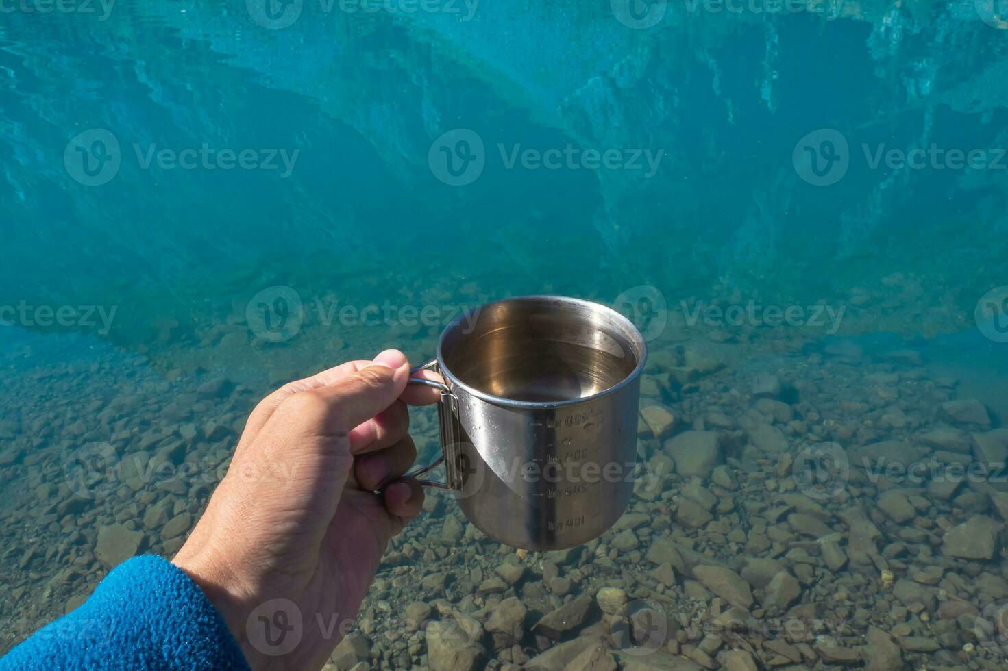Mug with pure glacial water of a mountain lake. Hand holding a hikers cup with clean drinking water. Clean drinking water in the blue mountain lake, close up. photo