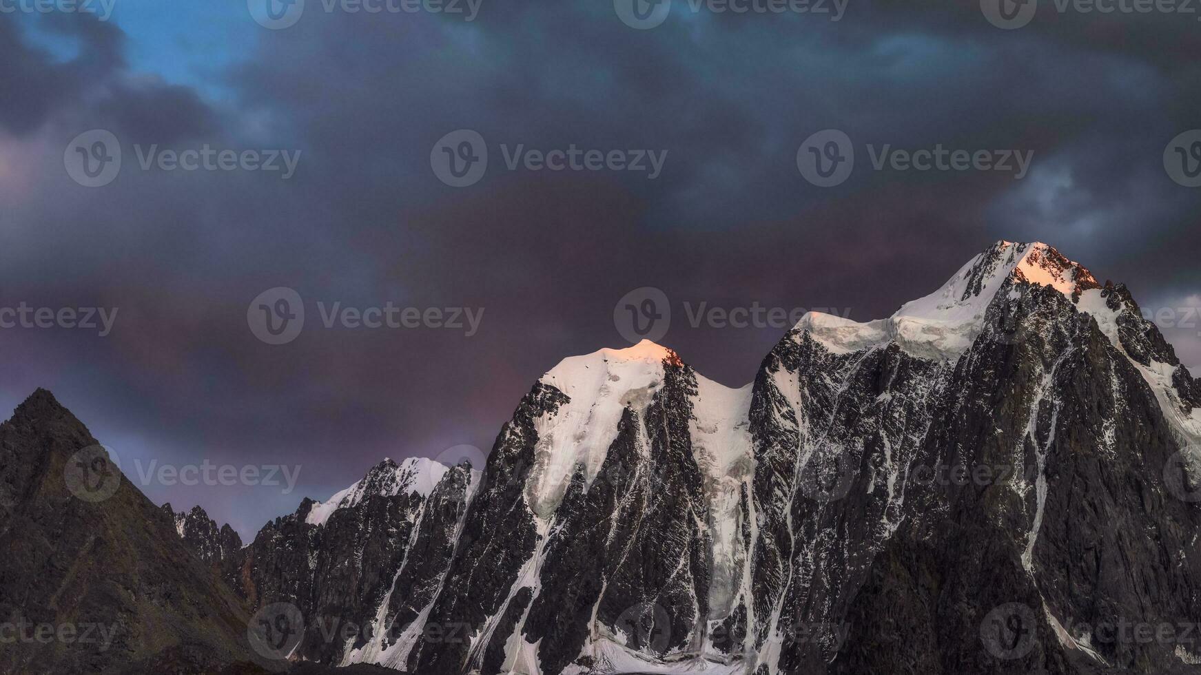 Darkness mountain landscape with great snowy mountain lit by dawn sun among dark clouds. Awesome alpine scenery with high mountain pinnacle at sunset or at sunrise. Big glacier on top in orange light. photo