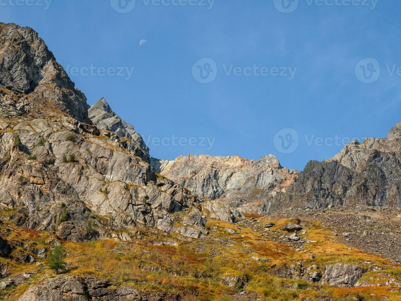 Bright mountain landscape with sharp rocks above yellow slope under blue sky with moon in sunny day. Colorful scenery with gold sunlit sharp rocky mountains. High rocky mountains in golden sunlight. photo