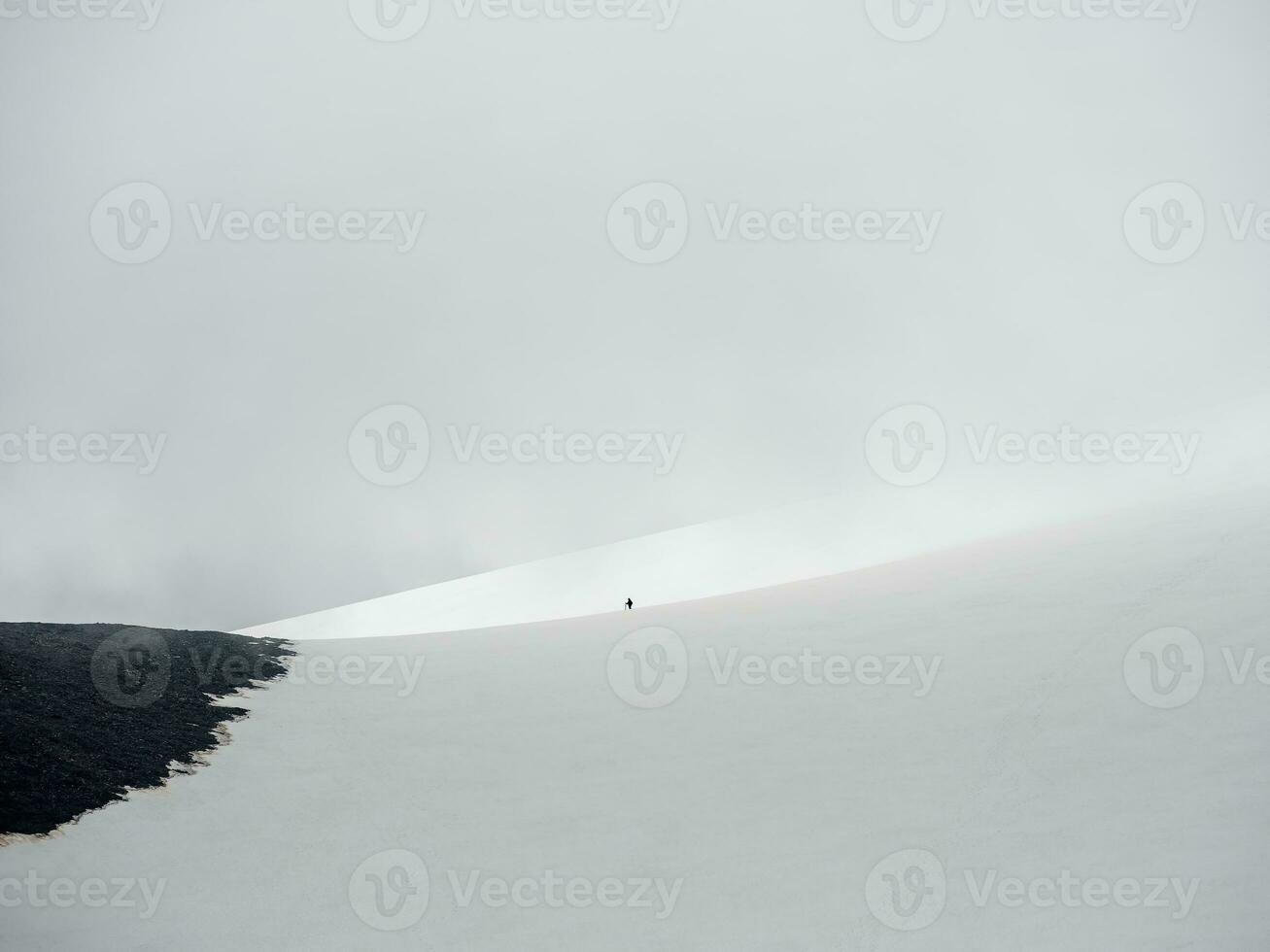 Minimalistic scenery with small man on large snowy mountain in sunlight under cloudy sky. Atmospheric landscape with sunshine on high snow mountain at changeable weather. Fog mountain in overcast. photo