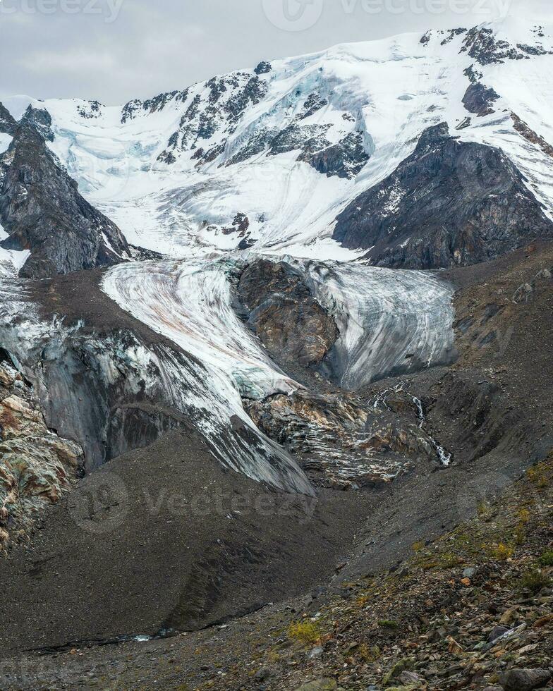 Vertical mountain landscape with long glacier tongue and dark snowy mountain wall. Scenic alpine landscape with glacial tongue and high mountain wall. photo