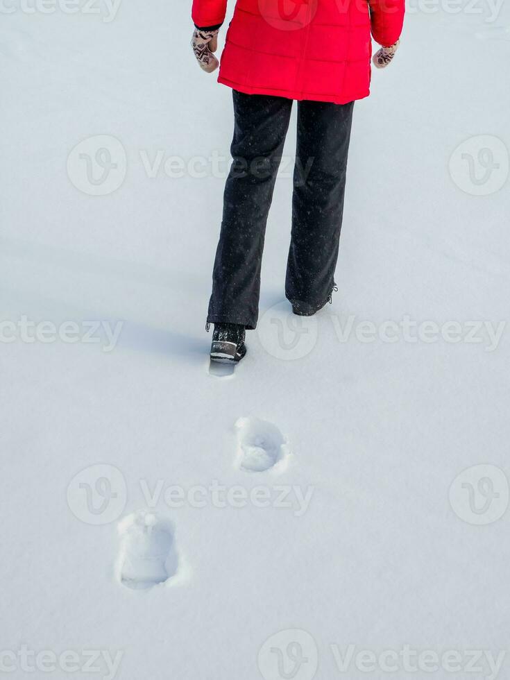 Woman in the red jacket walking on snow, footprints in snow, behind photo