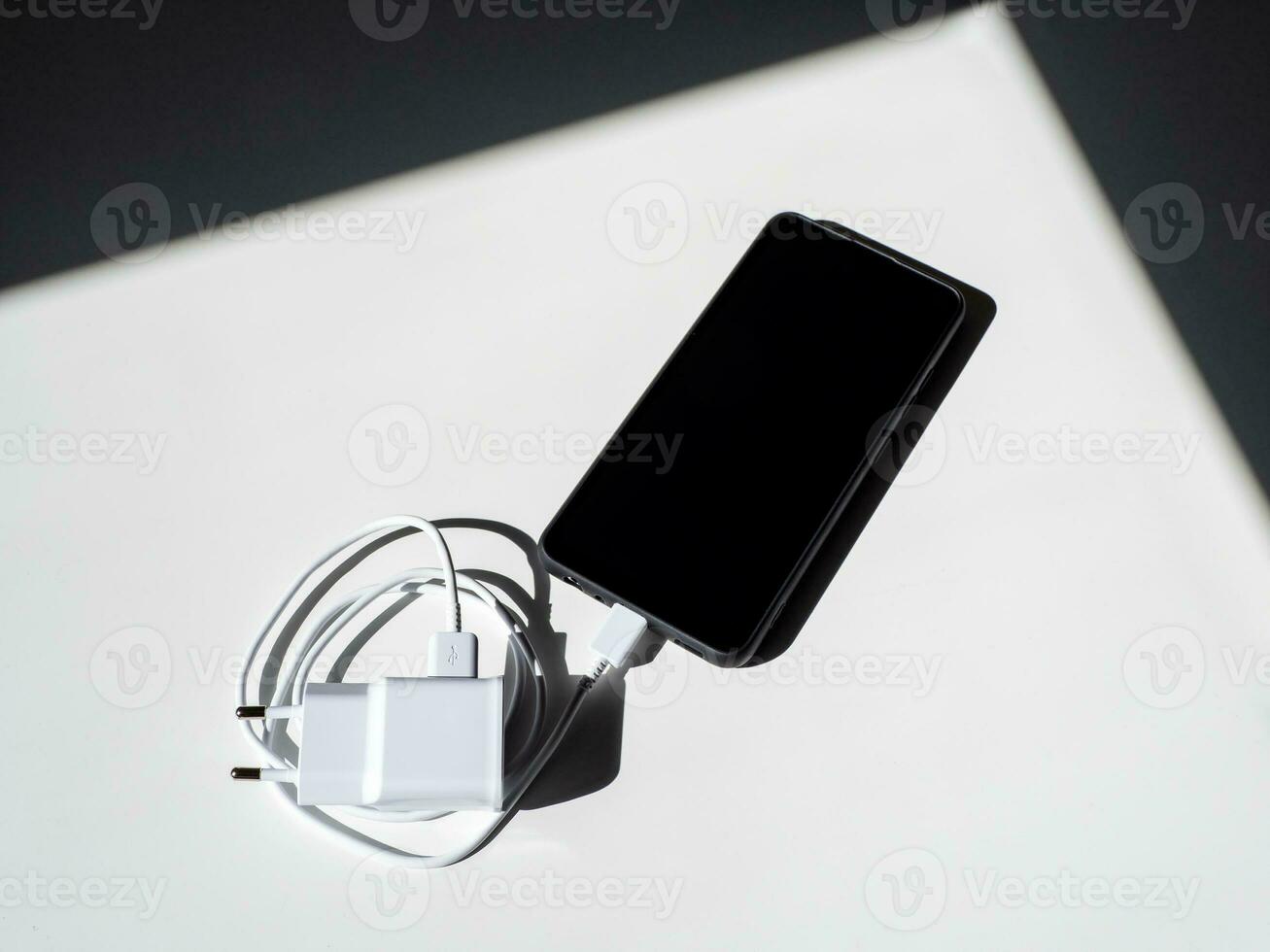 A black smartphone with a charging wire on a white table with shadows. photo