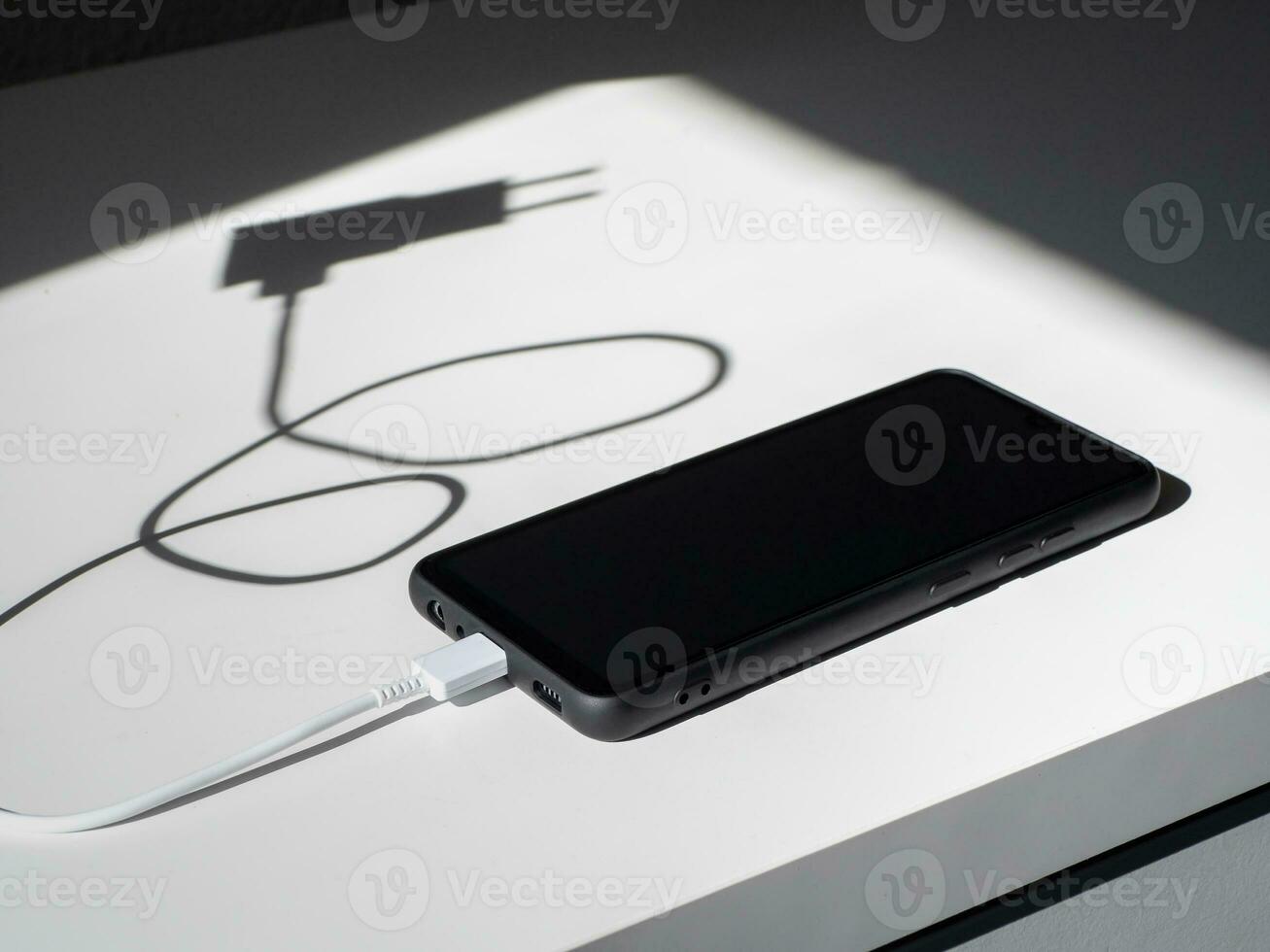 A black smartphone with a charging wire on a white table with shadows. photo