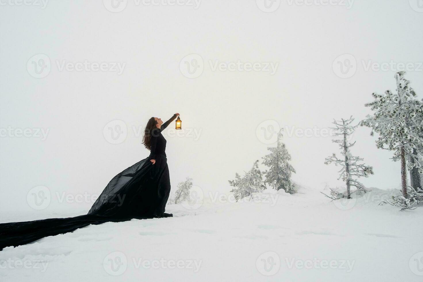 Young beautiful woman in long black dress with old lantern over winter hill background and snowfall. Fairy tale girl on polar winter landscape. Black witch in the snow. photo