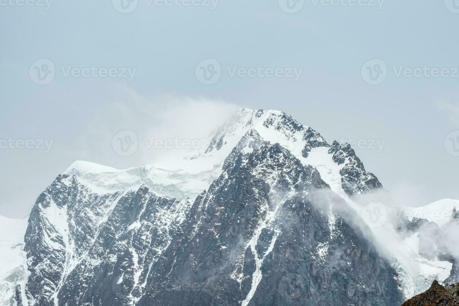 Winter atmospheric mountains landscape with blizzard in big snowy mountain top in low clouds. Awesome minimal scenery with white glacier on black rocks. High mountain pinnacle with snow in clouds. photo