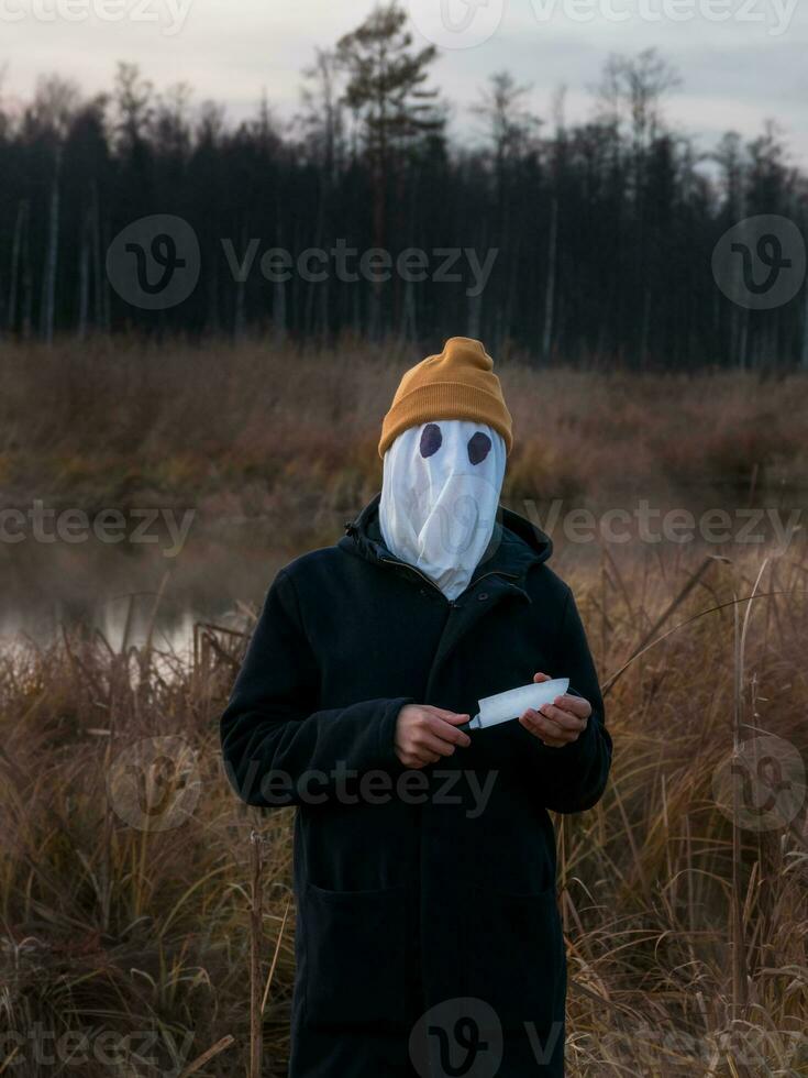 Faceless killer. Dangerous man with a white cloth on his face and a knife in his hand on the reeds in the evening. Halloween. photo