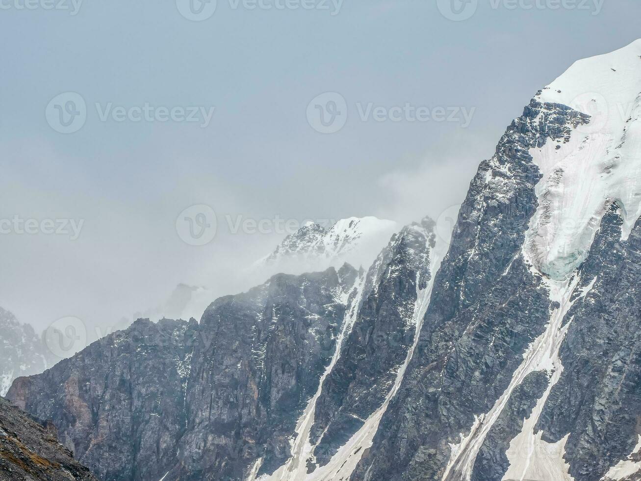 Winter atmospheric mountains landscape with blizzard in big snowy mountain top in low clouds. Awesome minimal scenery with white glacier on black rocks. High mountain pinnacle with snow in clouds. photo