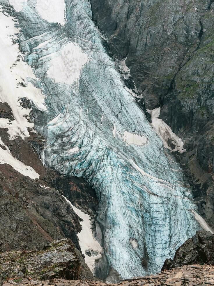 Vertical mountain landscape with blue long vertical glacier tongue with cracks among rocks. Aerial view to large glacier with icefall. Nature texture of mountain glacier with fissures close up. photo