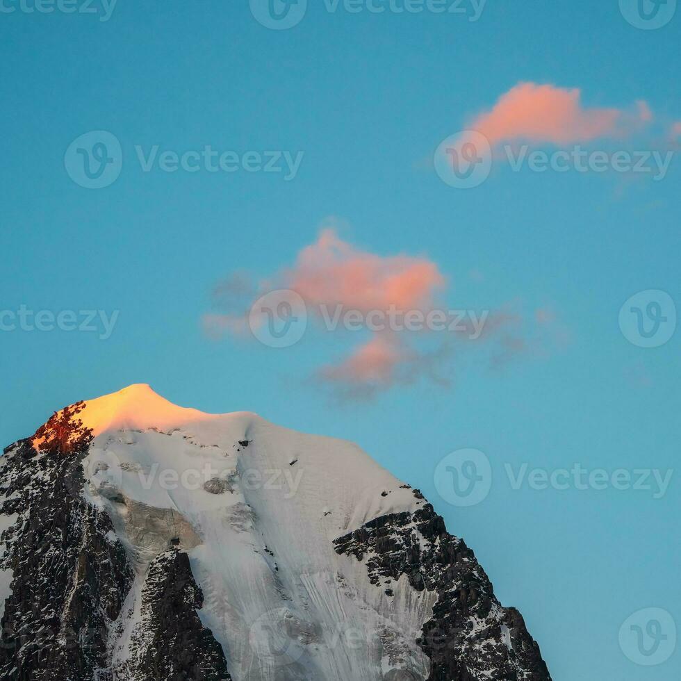 Top black rocks in evening golden sunshine and white-snow pointy peak. Atmospheric landscape with high snowy mountain with peaked top under cirrus clouds in sky. Square view. photo