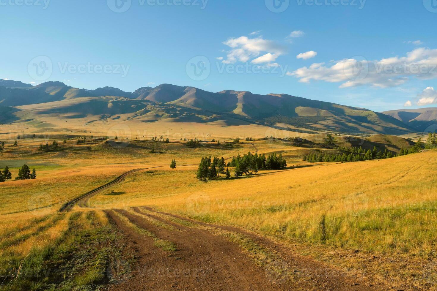 Beautiful golden mountain landscape with long dirt road through sunlit steppe to large mountains in white clouds on blue sky. Length road in big mountains. photo