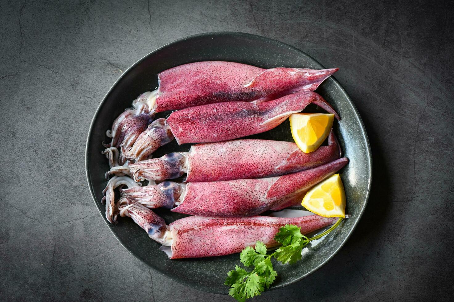 Raw squid on plate with salad spices lemon on the dark plate background, fresh squids octopus or cuttlefish for cooked food at restaurant or seafood market - top view photo