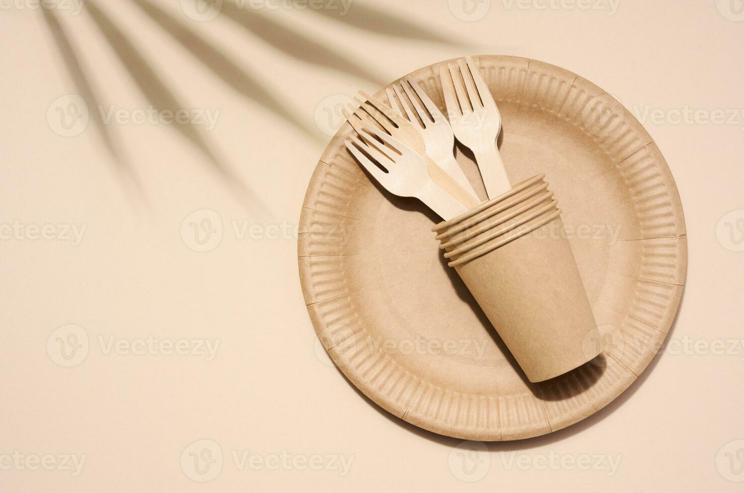 Brown paper plate, stack of glasses and wooden fork, disposable tableware on beige background, top view photo