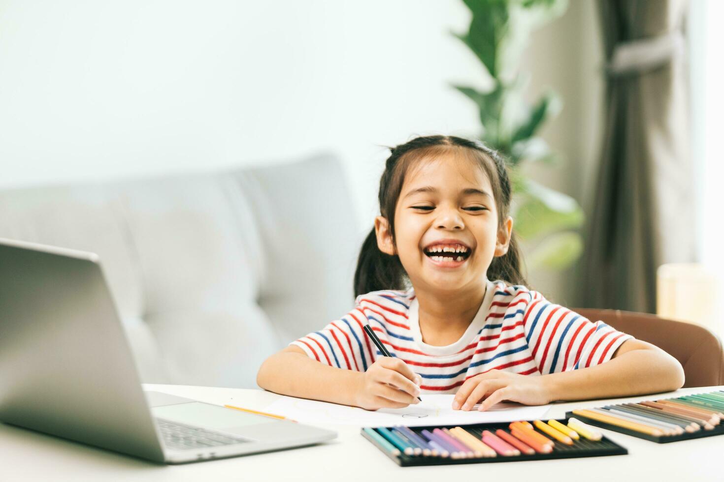 Little asian girl drawing with color pencils while using laptop at home photo