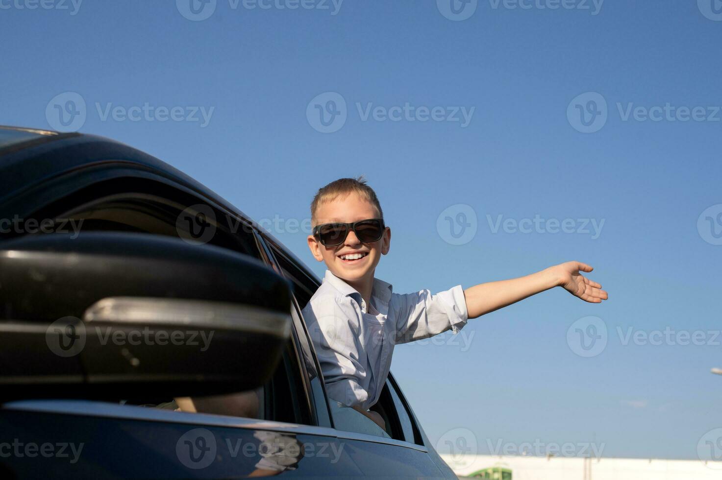 A cute boy in sunglasses looked out of the car window waving his hand with a laugh photo